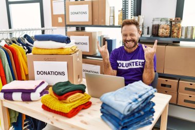 Middle age man wearing volunteer t shirt working with laptop shouting with crazy expression doing rock symbol with hands up. music star. heavy concept. 