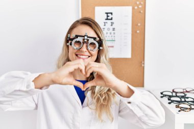Young beautiful optician woman wearing optometry glasses at the clinic smiling in love showing heart symbol and shape with hands. romantic concept. 