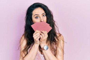 Young hispanic woman covering mouth with cards in shock face, looking skeptical and sarcastic, surprised with open mouth 