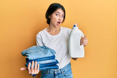 Young caucasian woman doing laundry holding detergent bottle and folded jeans afraid and shocked with surprise and amazed expression, fear and excited face. 