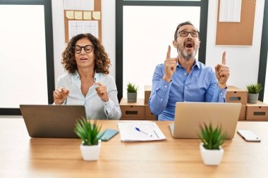 Middle age hispanic woman and man sitting with laptop at the office amazed and surprised looking up and pointing with fingers and raised arms. 