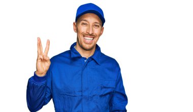 Bald man with beard wearing builder jumpsuit uniform showing and pointing up with fingers number three while smiling confident and happy. 