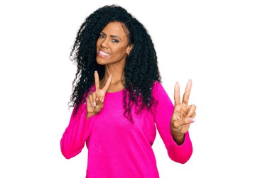 Orta yaşlı Afro-Amerikan kadın, günlük giysiler giymiş, gülümseyen parmaklarını zafer işareti yaparken gösteren kameraya bakıyor. İki numara.. 