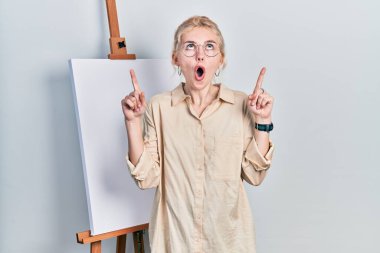 Beautiful caucasian woman with blond hair standing by painter easel stand amazed and surprised looking up and pointing with fingers and raised arms. 