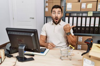 Handsome hispanic man working at small business commerce surprised pointing with hand finger to the side, open mouth amazed expression. 