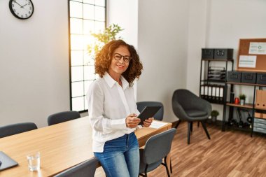 Orta yaşlı İspanyol kadın, ofiste dokunmatik ped kullanarak çalışırken kendine güvenerek gülümsüyor.