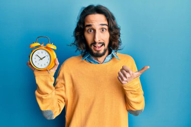 Çalar saati tutan genç İspanyol adam parmağıyla yan tarafı işaret ediyor. Ağzı açık gülümsüyor. 