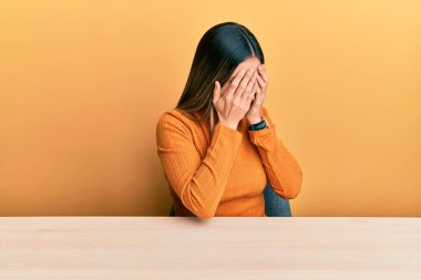 Young hispanic woman wearing casual clothes sitting on the table with sad expression covering face with hands while crying. depression concept. 