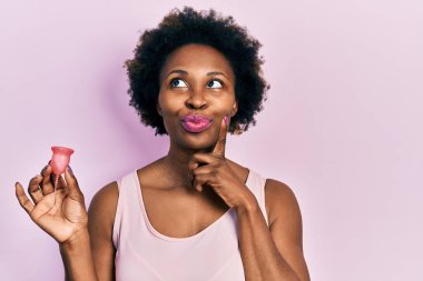 Young african american woman holding menstrual cup serious face thinking about question with hand on chin, thoughtful about confusing idea 