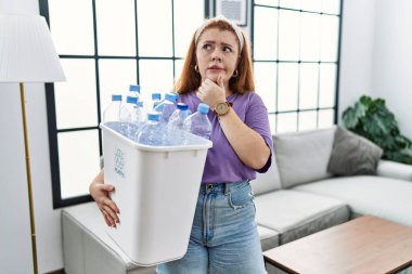 Elinde plastik şişelerle çöpleri geri dönüşüm sepetini tutan genç kızıl saçlı kadın bir soru hakkında endişeli ve gergin bir şekilde düşünüyor. 