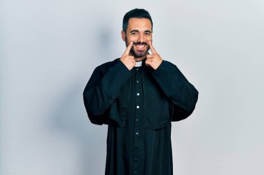 Handsome hispanic man with beard wearing catholic priest robe smiling with open mouth, fingers pointing and forcing cheerful smile 