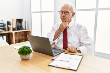 Senior man working at the office using computer laptop thinking concentrated about doubt with finger on chin and looking up wondering 