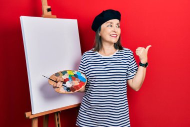 Orta yaşlı, İspanyol bir kadın elinde paletle ayakta dikiliyor. Resim sehpası ayakta gülümsüyor, gülümsüyor ve baş parmağıyla yan tarafa bakıyor.. 