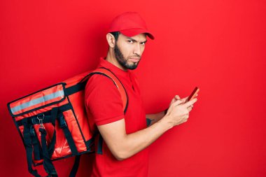 Hispanic man with beard wearing delivery package backpack using smartphone skeptic and nervous, frowning upset because of problem. negative person. 