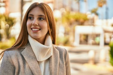 Genç kızıl kız gülümsüyor, mutlu bir şekilde şehirde duruyor..