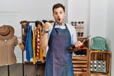 Handsome young man working as manager at retail boutique surprised pointing with hand finger to the side, open mouth amazed expression. 