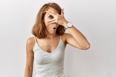 Beautiful caucasian woman standing over isolated background peeking in shock covering face and eyes with hand, looking through fingers with embarrassed expression. 