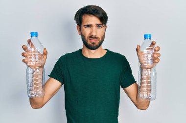 Young hispanic man holding recycling plastic bottles clueless and confused expression. doubt concept. 