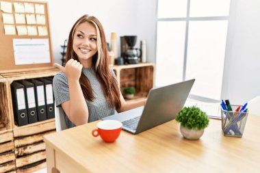 Young brunette woman working at the office with laptop smiling with happy face looking and pointing to the side with thumb up. 