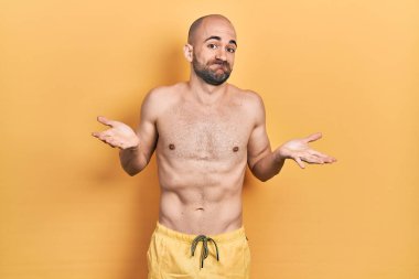 Young bald man wearing swimwear clueless and confused expression with arms and hands raised. doubt concept. 