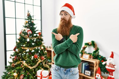 Redhead man with long beard wearing christmas hat by christmas tree pointing to both sides with fingers, different direction disagree 