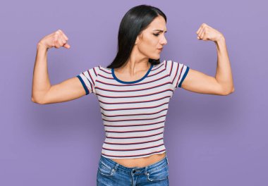 Young hispanic girl wearing casual striped t shirt showing arms muscles smiling proud. fitness concept. 