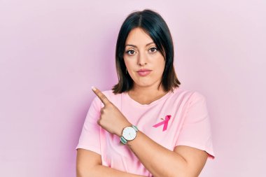 Young hispanic woman wearing pink cancer ribbon on t shirt pointing with hand finger to the side showing advertisement, serious and calm face 
