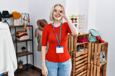 Young caucasian woman working as manager at retail boutique doing ok gesture with hand smiling, eye looking through fingers with happy face. 