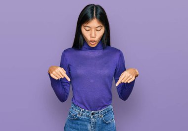 Beautiful young asian woman wearing casual clothes pointing down with fingers showing advertisement, surprised face and open mouth 