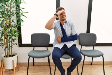 Handsome young man sitting at doctor waiting room with arm injury very happy and smiling looking far away with hand over head. searching concept. 