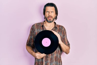 Middle age caucasian man holding vinyl disc in shock face, looking skeptical and sarcastic, surprised with open mouth 