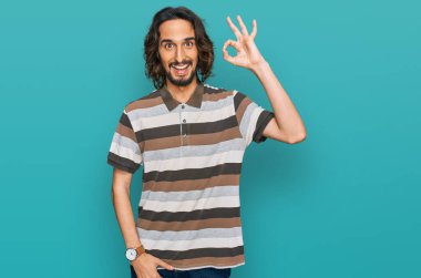 Gündelik kıyafetler giyen genç İspanyol adam gülümsüyor. El ve parmaklarıyla 