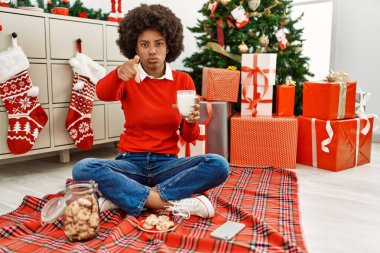 Young african american woman with afro hair eating pastries sitting by christmas tree pointing with finger to the camera and to you, confident gesture looking serious 