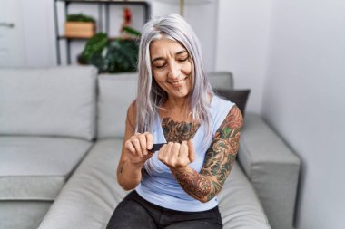 Orta yaşlı, kır saçlı, tırnak kireci kullanan kadın evdeki kanepede oturuyor.