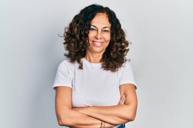 Orta yaşlı, kollarını kavuşturup gülümseyen, yüzünde mutlu ve havalı bir gülümsemeyle İspanyol bir kadın. diş gösterme. 