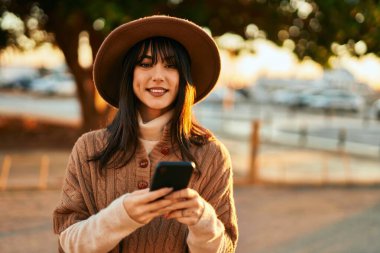 Kışlık şapkalı esmer kadın gün batımında şehrin dışında akıllı telefon kullanarak gülümsüyor.