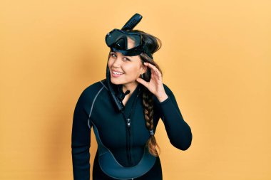 Young hispanic girl wearing diver neoprene uniform smiling with hand over ear listening and hearing to rumor or gossip. deafness concept. 