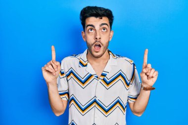 Gündelik giysiler giyen genç İspanyol adam şaşırmış ve şaşırmış bir şekilde yukarı bakıyor ve parmaklarıyla işaret ediyor ve kollarını kaldırıyor.. 