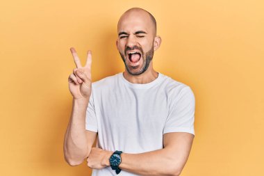 Young bald man wearing casual white t shirt smiling with happy face winking at the camera doing victory sign with fingers. number two. 