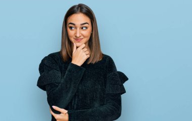 Beautiful brunette woman wearing elegant sweater with hand on chin thinking about question, pensive expression. smiling with thoughtful face. doubt concept. 