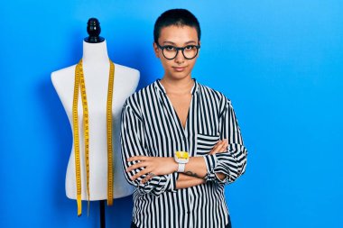 Beautiful hispanic woman with short hair standing by manikin with crossed arms relaxed with serious expression on face. simple and natural looking at the camera. 