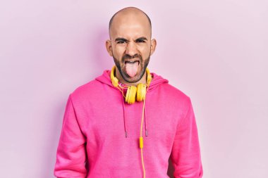 Young bald man wearing gym clothes and using headphones sticking tongue out happy with funny expression. emotion concept. 