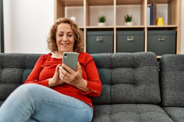 Orta yaşlı beyaz kadın akıllı telefon kullanıyor. Evdeki kanepede oturuyor..