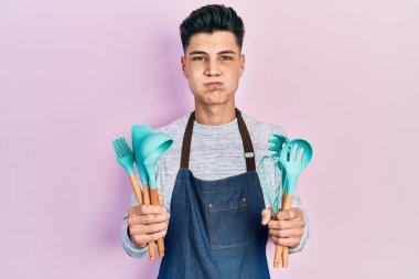 Young hispanic man wearing professional baker apron holding cooking tools puffing cheeks with funny face. mouth inflated with air, catching air. 
