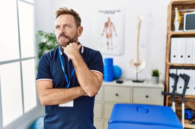 Middle age physiotherapist man working at pain recovery clinic with hand on chin thinking about question, pensive expression. smiling with thoughtful face. doubt concept. 