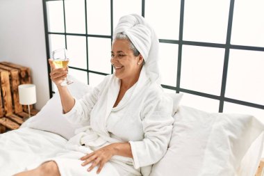 Middle age grey-haired woman drinking champagne sitting on the bed at bedroom.