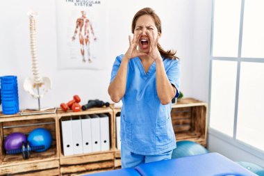 Middle age hispanic physiotherapist woman working at pain recovery clinic shouting angry out loud with hands over mouth 