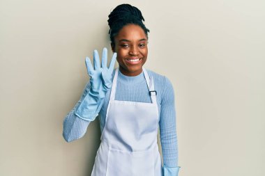African american woman with braided hair wearing cleaner apron and gloves showing and pointing up with fingers number four while smiling confident and happy. 