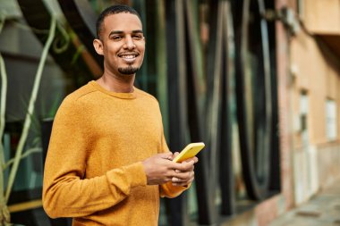 Genç Afrikalı Amerikalı adam şehirde akıllı telefon kullanarak mutlu bir şekilde gülümsüyor..