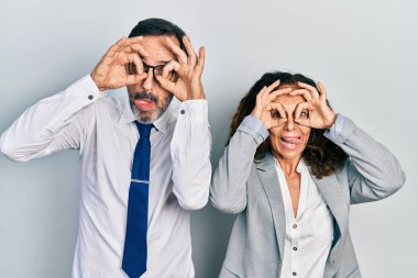Middle age couple of hispanic woman and man wearing business office uniform doing ok gesture like binoculars sticking tongue out, eyes looking through fingers. crazy expression. 